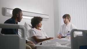 Young black male in hospital bed with parental figure to the left and medical provider to the right
