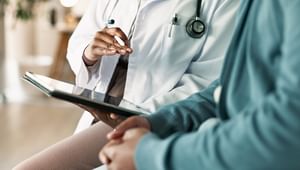 Close up of a medical professional's hands and a tablet sitting next to a patient with their hands crossed on their lap