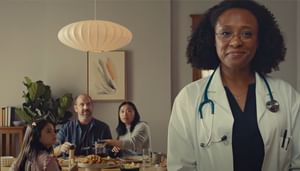 Doctor to the right with family looking at her while sitting at the table for dinner.