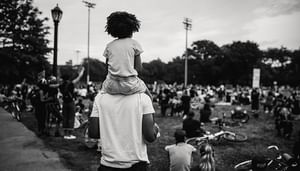 Young son on fathers back from behind in field with people sitting on grass in black and white